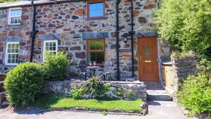 A stone house with a wooden door, two windows, and a small outdoor seating area with a table and chairs.