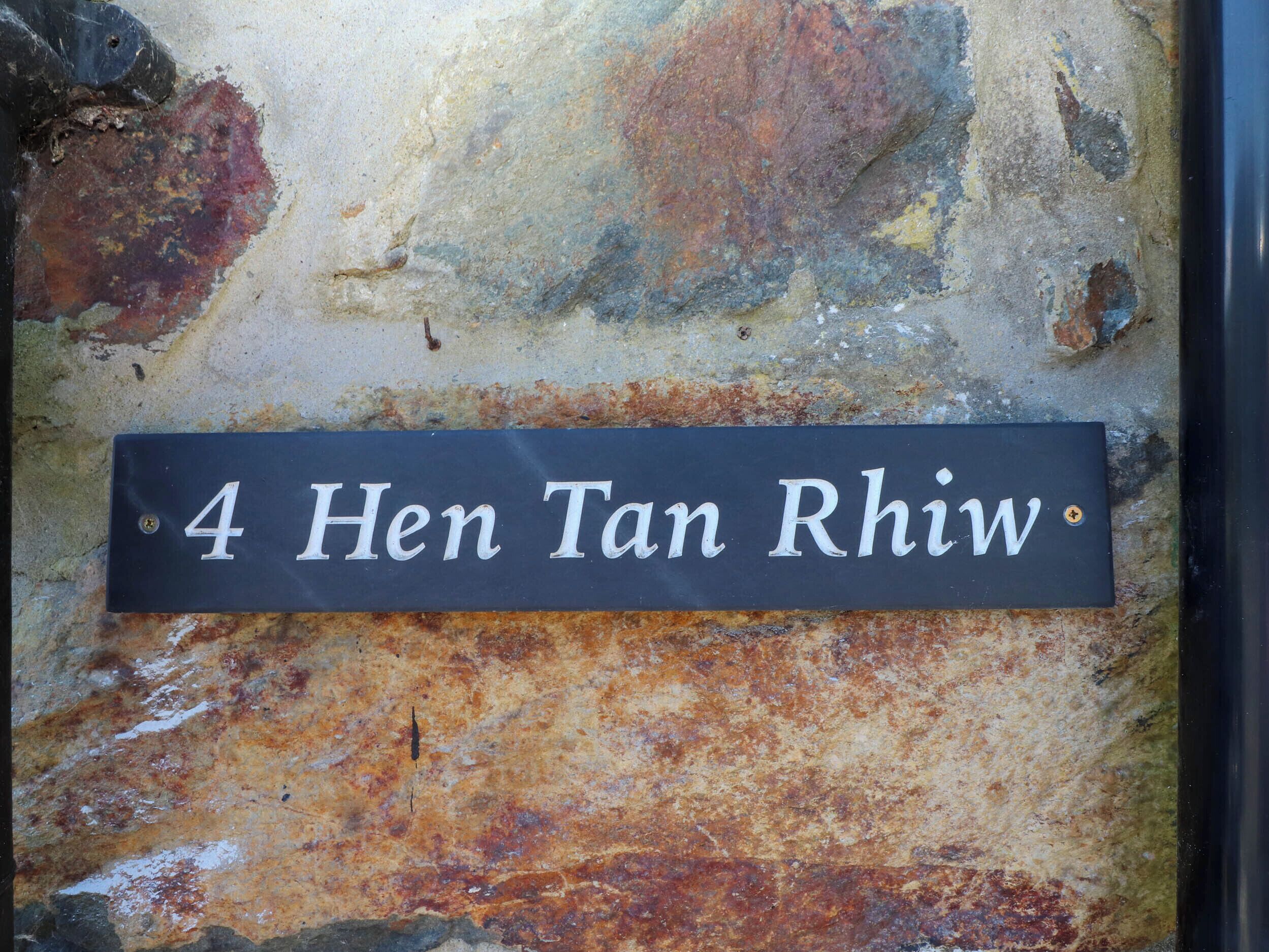 A house number sign with "4 Hen Tan RhIw" on a black background, mounted on a textured wall with visible rust and discoloration.