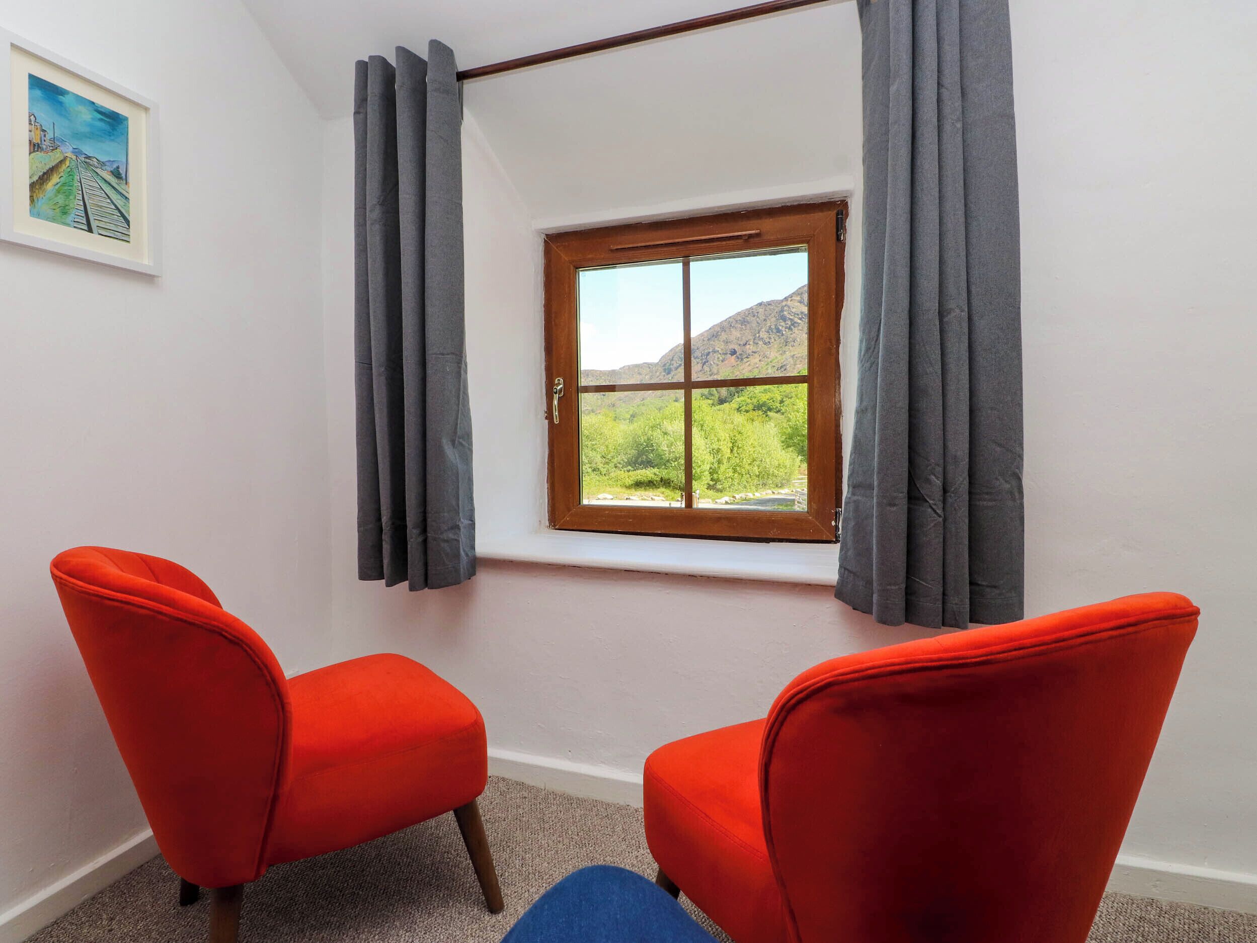 A room with two red chairs, a window with a view of greenery and mountains, and a framed painting on the wall.