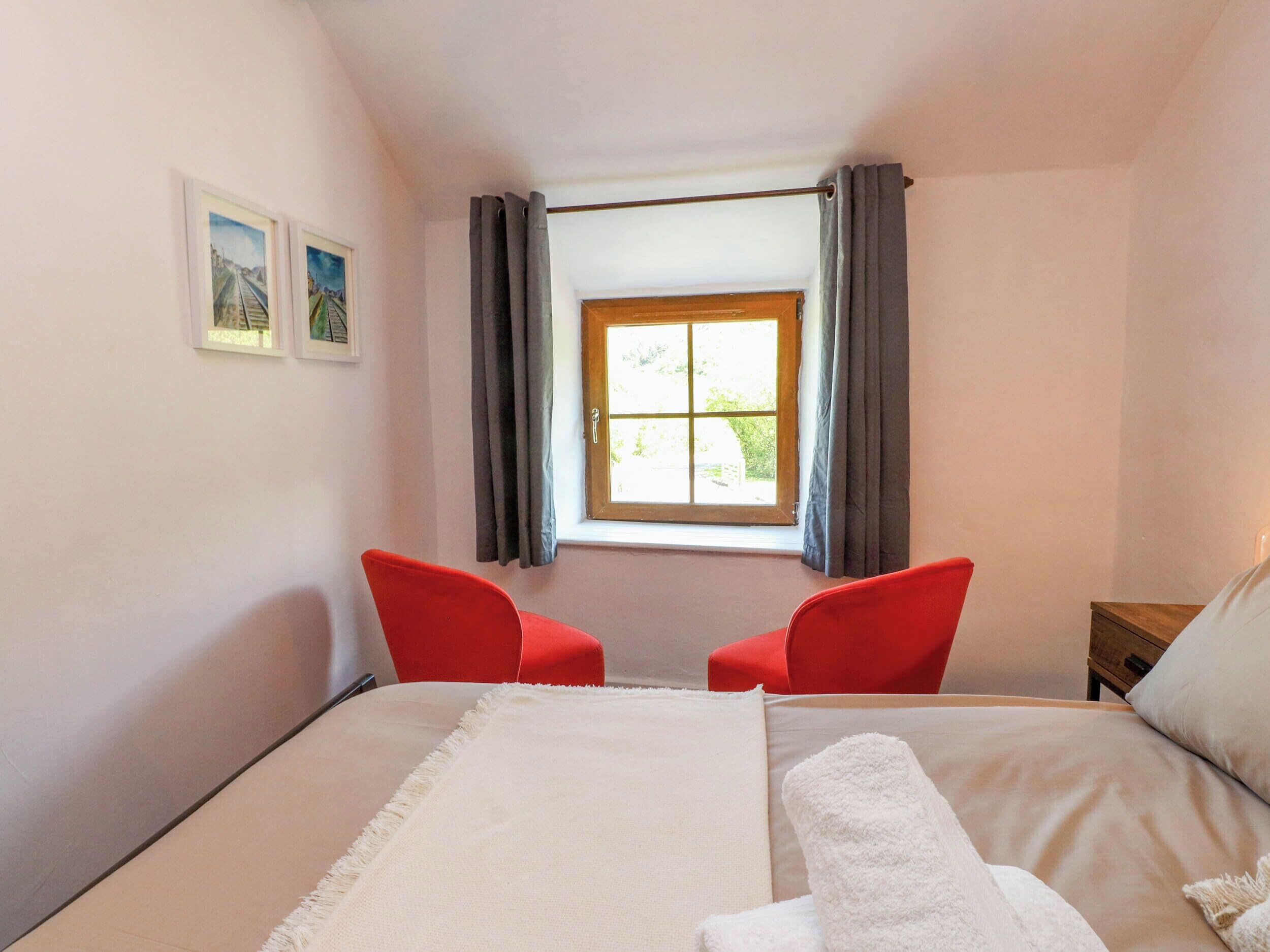 A bedroom with a bed, two red chairs, a window with curtains, and two framed pictures on the wall.