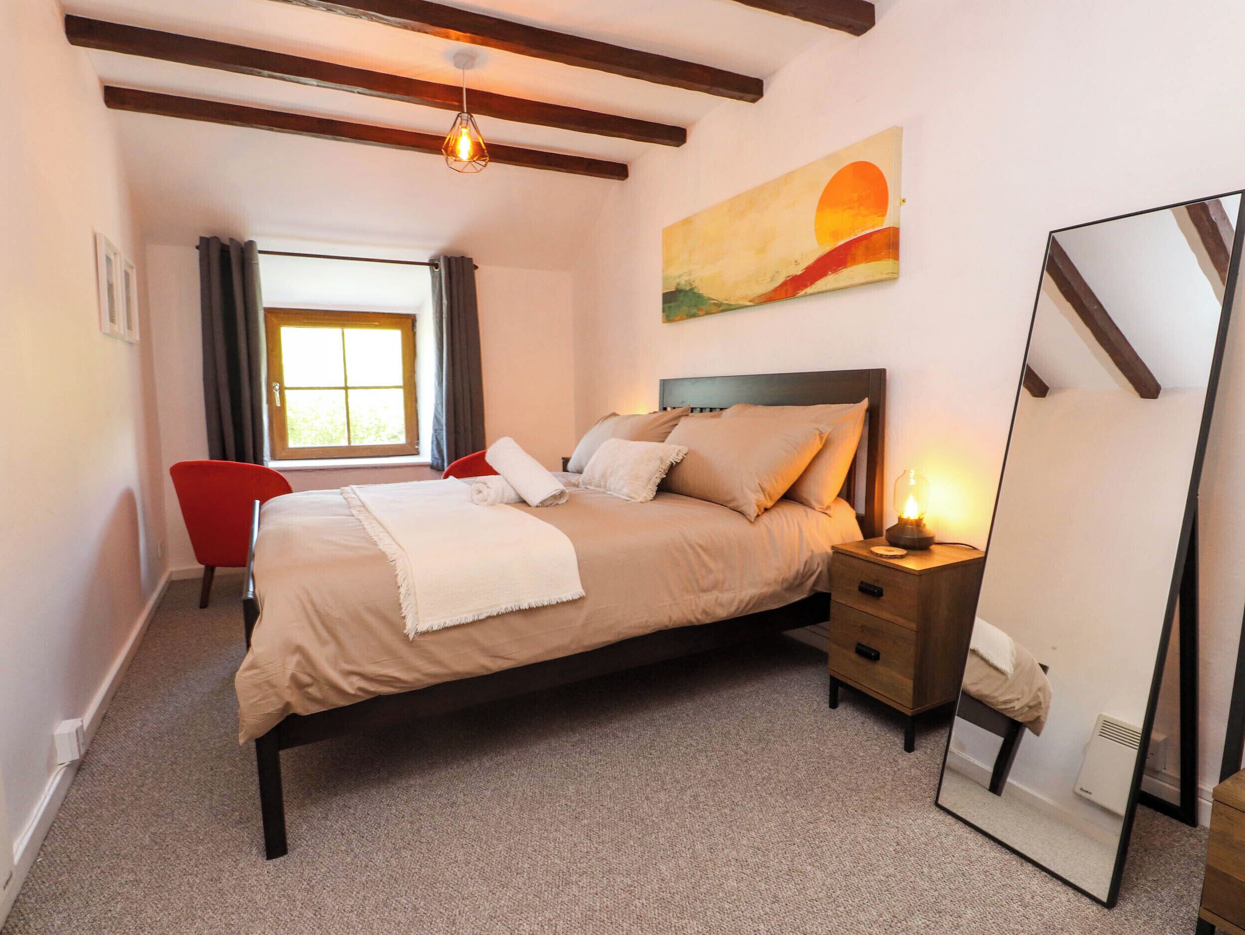 A bedroom with a bed, bedside table, lamp, chair, window, and artwork.
