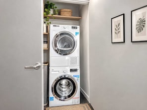 A laundry room with stacked washing machines, a shelf with plants, and framed botanical prints on the wall. - Stylish 2BR | Heart of Town | Host 16 | Valet Park (Dallas)