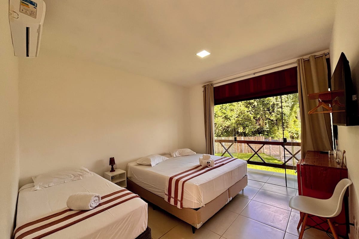 A hotel room with two beds, a desk, and a chair. There is a window with a view of greenery outside.