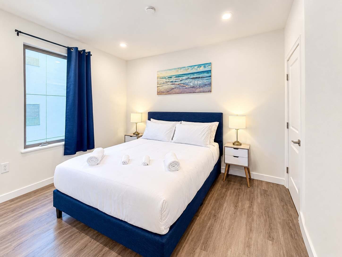 Una habitación de hotel con una cama grande, dos mesitas de noche con lámparas, un cuadro en la pared y una ventana con cortinas.