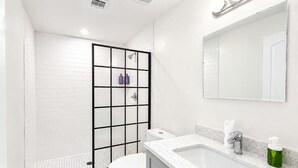 Een moderne badkamer met een witte betegelde douche, toilet, wastafel met spiegel en een groene zeepdispenser.