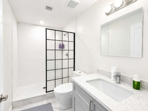 A modern bathroom with a white tiled shower, a toilet, a sink with a mirror, and a green soap dispenser.