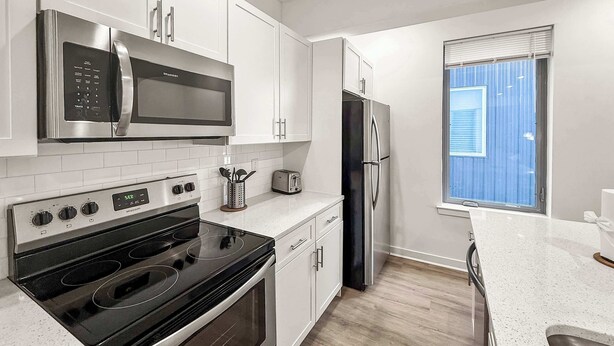 A modern kitchen with white cabinets, stainless steel appliances, a black stove, and a window with blinds. - Ideal 2BD Retreat | Sleeps 6 | Center City Philly (Philadelphia)