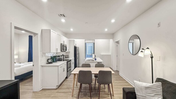 A modern kitchen with white cabinets, a dining table with chairs, a black sofa with a pillow, and a floor lamp.