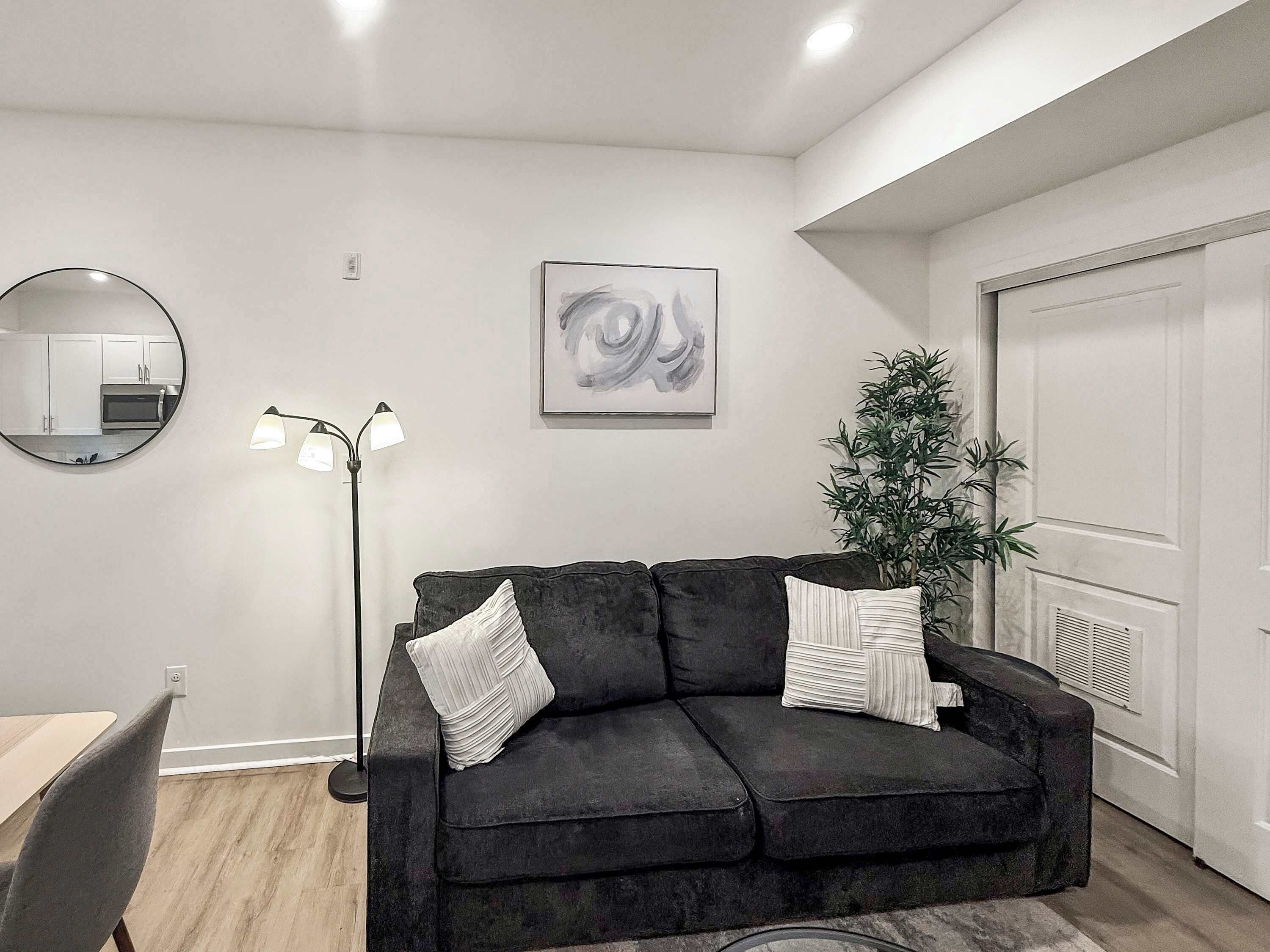 Una sala de estar moderna con un sofá gris oscuro, un espejo redondo, una lámpara de pie, un cuadro abstracto enmarcado y una planta en maceta.