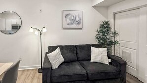 A modern living room with a dark grey sofa, a round mirror, a floor lamp, a framed abstract painting, and a potted plant.