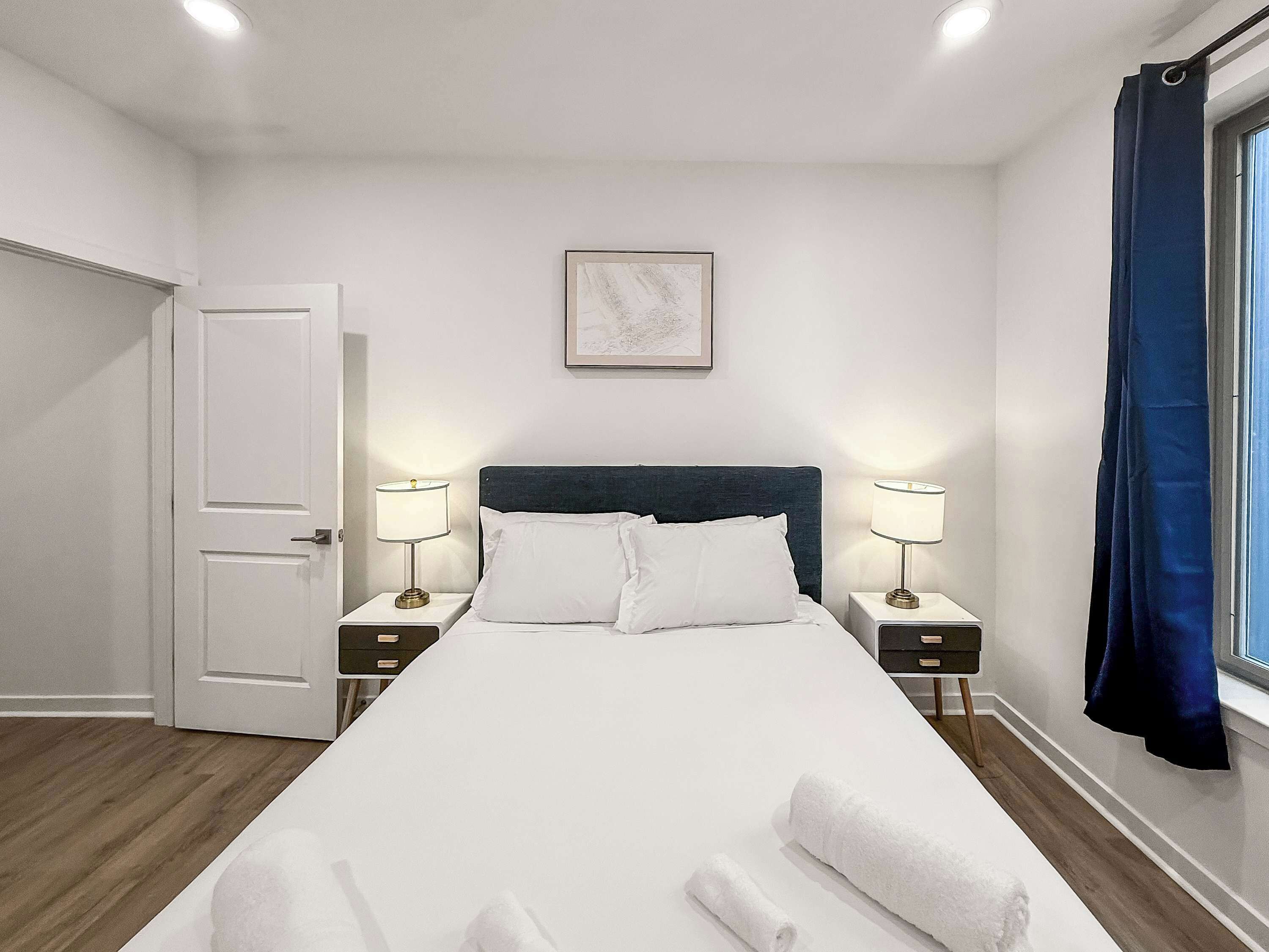 A neatly made bed with white linens, two bedside tables with lamps, a door, a framed picture on the wall, and a window with a blue curtain.