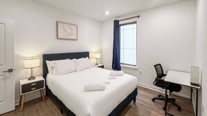 A hotel room with a bed, bedside tables, a desk with a chair, a window with curtains, and a wall-mounted picture. - Ideal 2BD Retreat | Sleeps 6 | Center City Philly (Philadelphia)