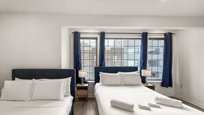 A hotel room with two beds, a window with curtains, a nightstand, and a view of buildings.