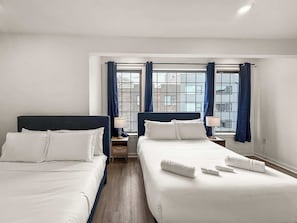 A hotel room with two beds, a window with curtains, a nightstand, and a view of buildings.