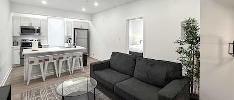 Una sala de estar moderna con un sofá gris, una mesa de centro de vidrio y una zona de cocina con armarios y electrodomésticos blancos.