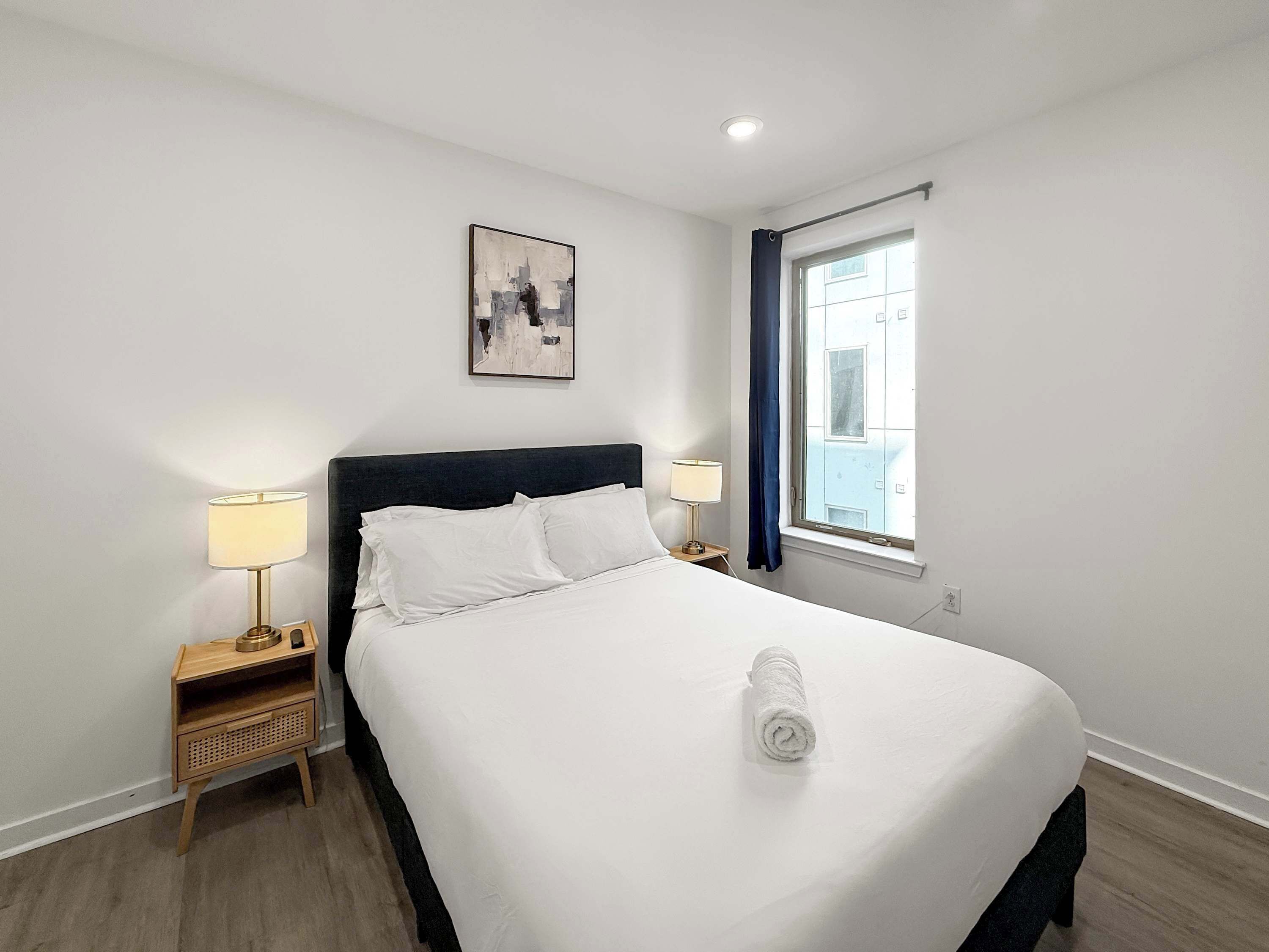 Une chambre d’hôtel avec un lit, deux tables de chevet avec des lampes, une fenêtre avec des rideaux, un tableau au mur et un plancher en bois.