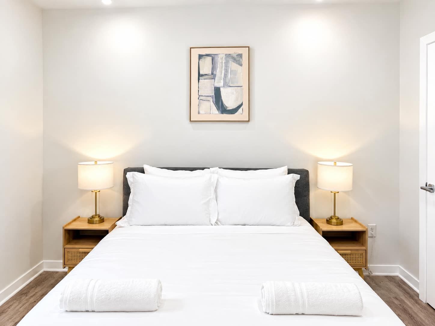 Una habitación de hotel con una cama, dos lámparas de noche, un cuadro abstracto enmarcado y una puerta.