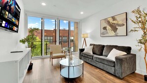 Una sala de estar moderna con sofá, sillón y mesa de centro, ventanas amplias con vista a edificios, y un televisor montado que muestra varios canales.
