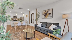 A modern living room with a grey sofa, a round glass coffee table, and a dining area with white chairs.
