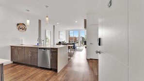 A modern kitchen with a dishwasher, a dining area with a table and chairs, and a view of the cityscape through large windows.