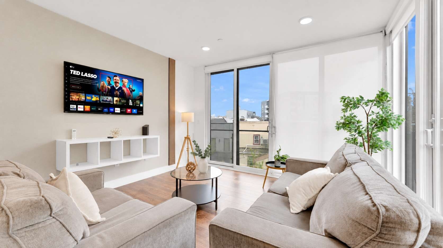A modern living room with a flat-screen TV, sofas, a coffee table, and a view of the city through large windows.