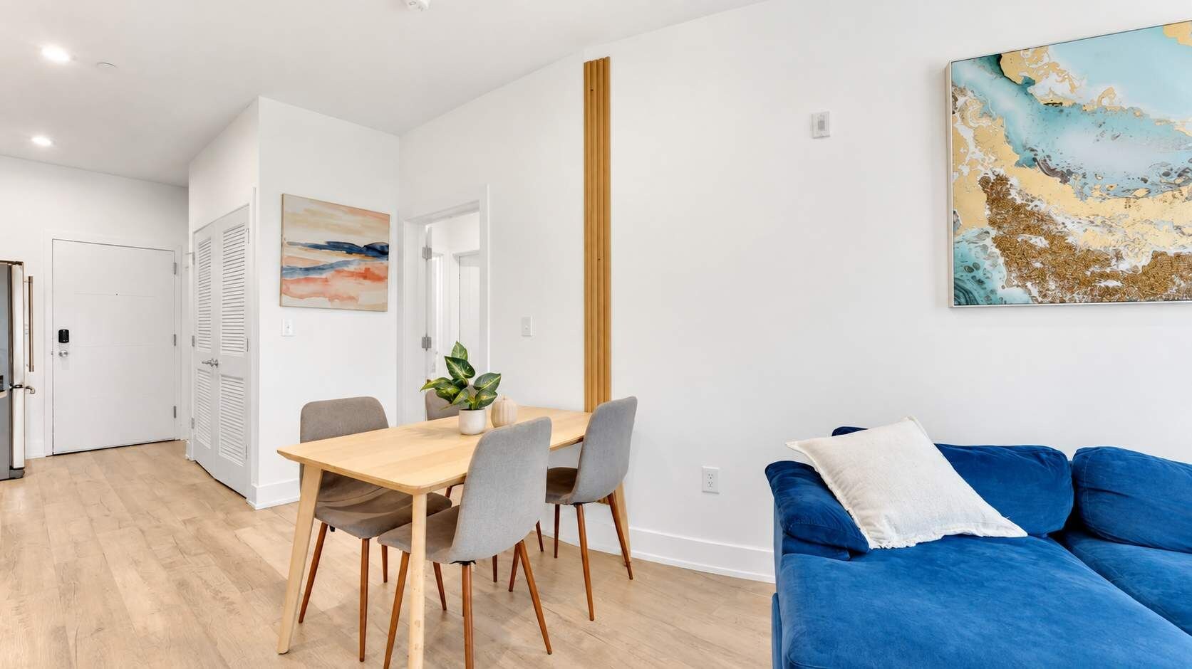 A modern living room with a blue sofa, a wooden dining table with chairs, and a painting on the wall.