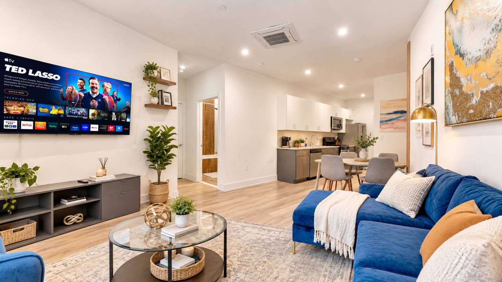 Una sala de estar moderna con una televisión de pantalla plana, un sofá azul, una mesa de centro de vidrio y una cocina al fondo.