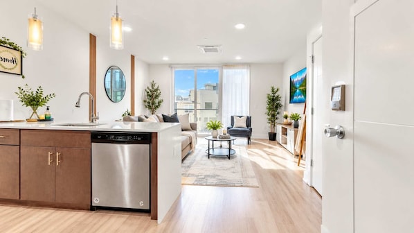 A modern kitchen with a dishwasher, a dining area with a round table, and a view of the city through large windows.