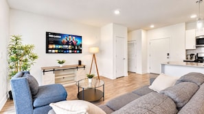 A modern living room with a sofa, armchair, and a coffee table. There is a flat-screen TV mounted on the wall displaying a sports channel.