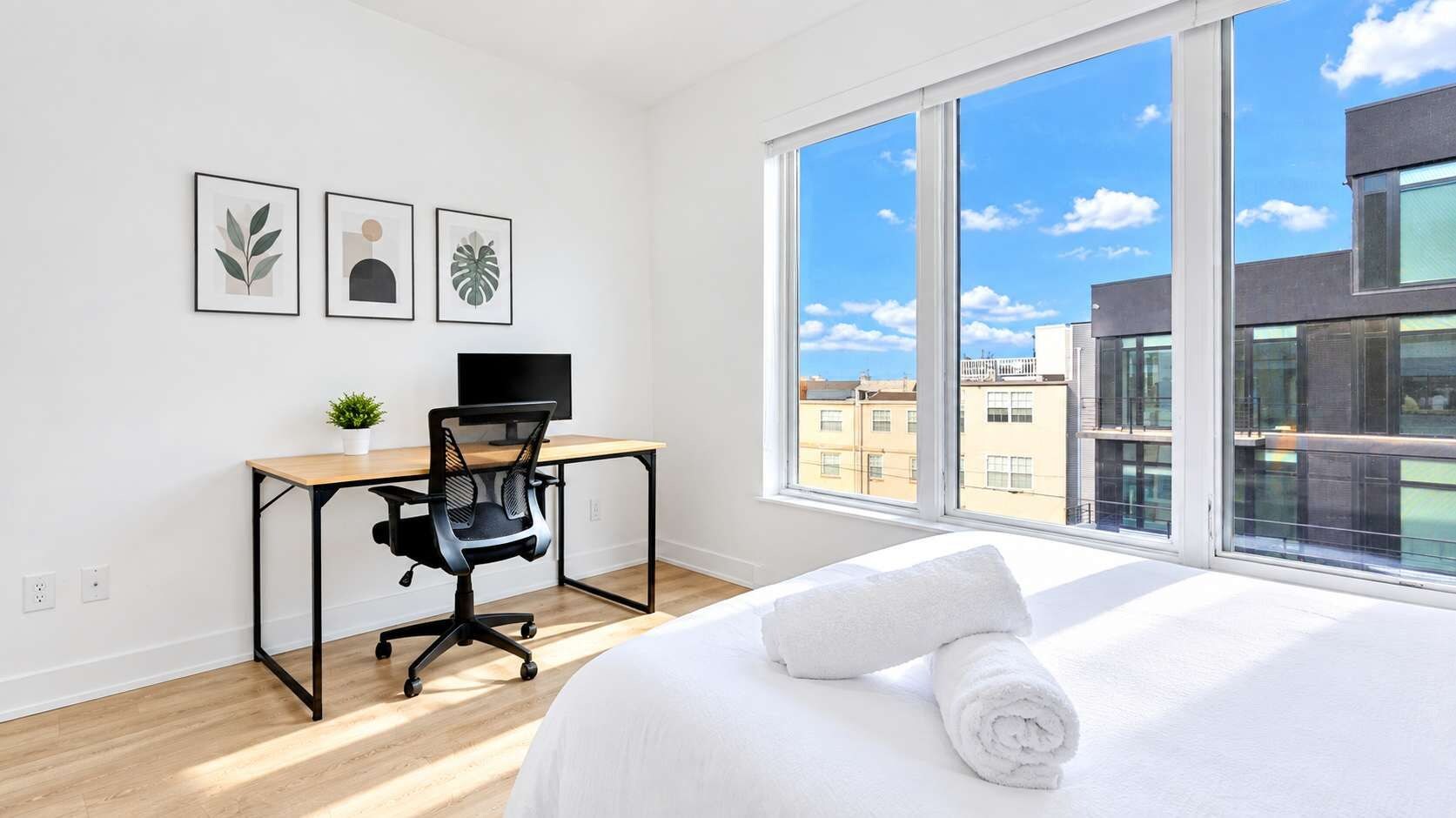 A hotel room with a large window, a bed with white linens, a desk with a computer, and framed artwork on the wall.