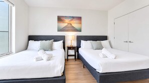 Une chambre d’hôtel avec deux lits, une table de chevet avec une lampe, un tableau représentant un coucher de soleil sur une jetée et une armoire.