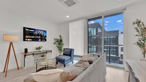 Una sala de estar moderna con un sofá, una silla, una mesa de centro, un televisor y un amplio ventanal con vistas a edificios.