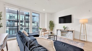 A modern living room with a sofa, a coffee table, a TV mounted on the wall, and a view of a building through large windows.