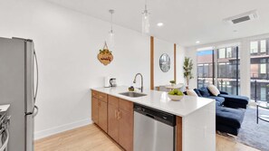 Una cocina moderna con lavavajillas, un fregadero y una encimera. Hay una zona de comedor con una mesa y sillas, y un sofá oscuro.