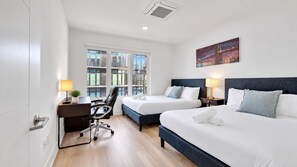 A hotel room with two beds, a desk, a chair, and a large window with a view of buildings.