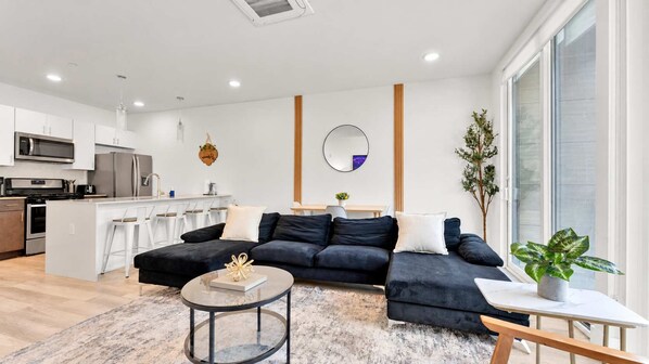 A modern living room with a dark sofa, a round coffee table, and a kitchen in the background.