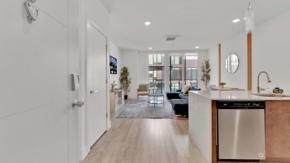 A modern kitchen with a dishwasher, a dining area with a table and chairs, and a living room with a sofa and a TV.