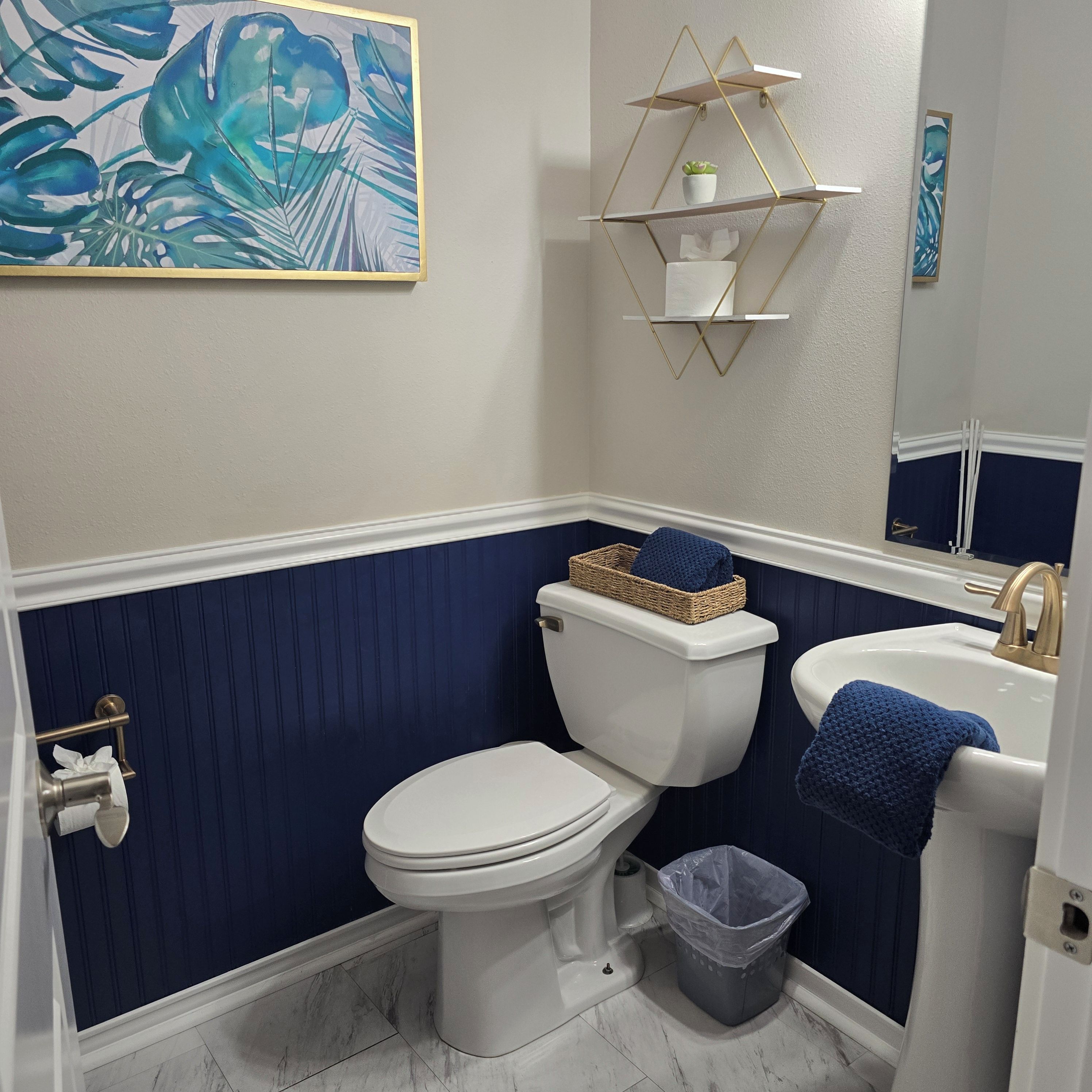 Une salle de bain avec des toilettes blanches, un lavabo, une poubelle et une étagère décorative agrémentée d’une plante.
