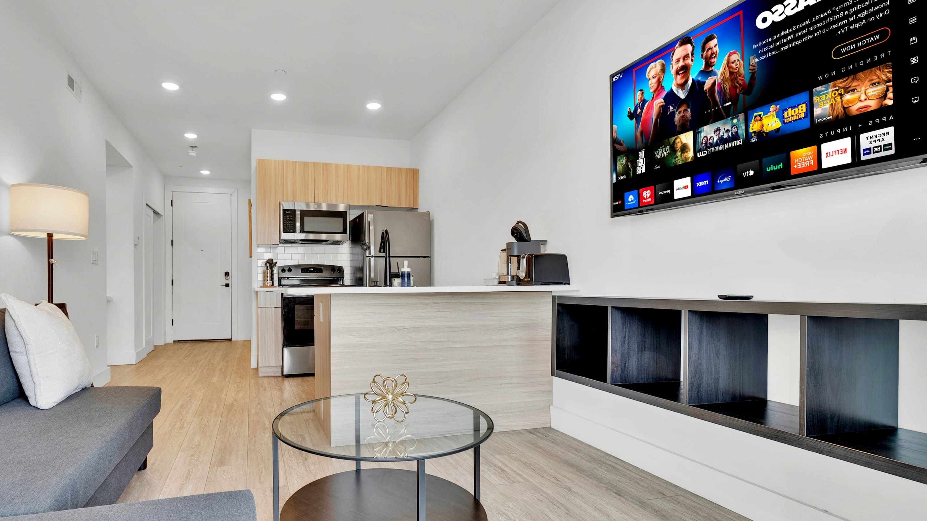 Een moderne woonkamer met een flatscreen televisie aan de muur, een glazen salontafel en een grijze bank.