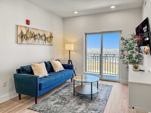 A living room with a blue sofa, a glass coffee table, a lamp, a painting, and a balcony with a view.