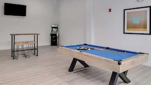 Ein Billardtisch in einem Raum mit Holzboden, einem kleinen Tisch mit Hocker, einem Automaten und einem gerahmten Bild an der Wand.