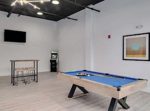 A pool table in a room with a wooden floor, a small table with stools, a vending machine, and a framed picture on the wall.