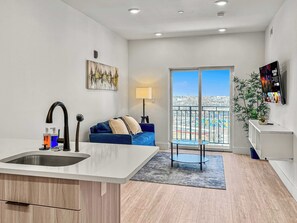 A modern kitchen with a central island, a dining area with a blue sofa, a large window with a city view, and a TV mounted on the wall.