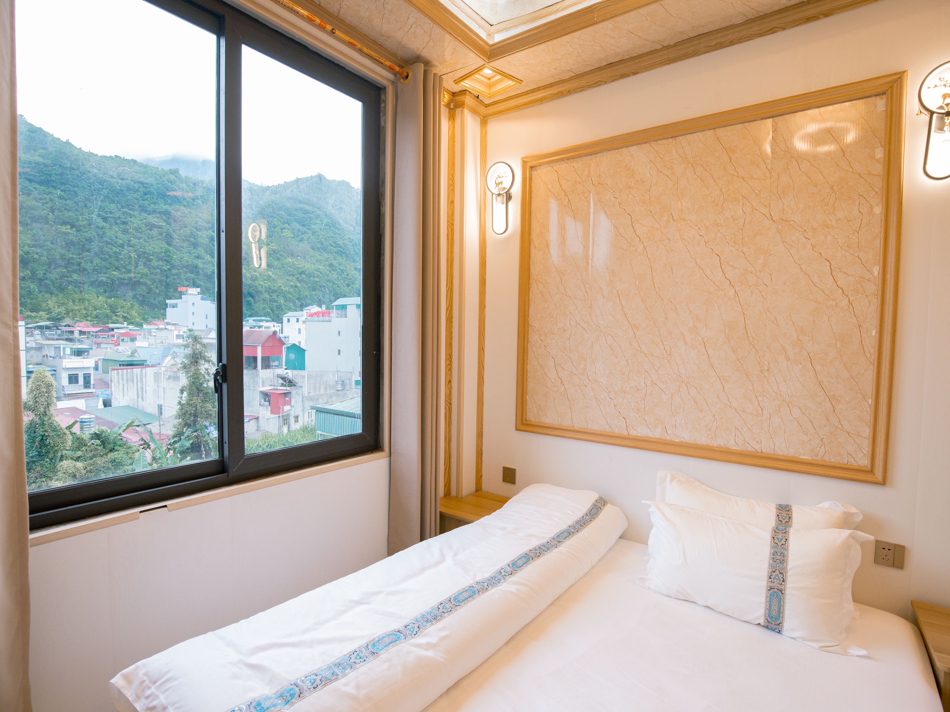 A hotel room with a bed, a window with a view of buildings and mountains, and a marble wall.
