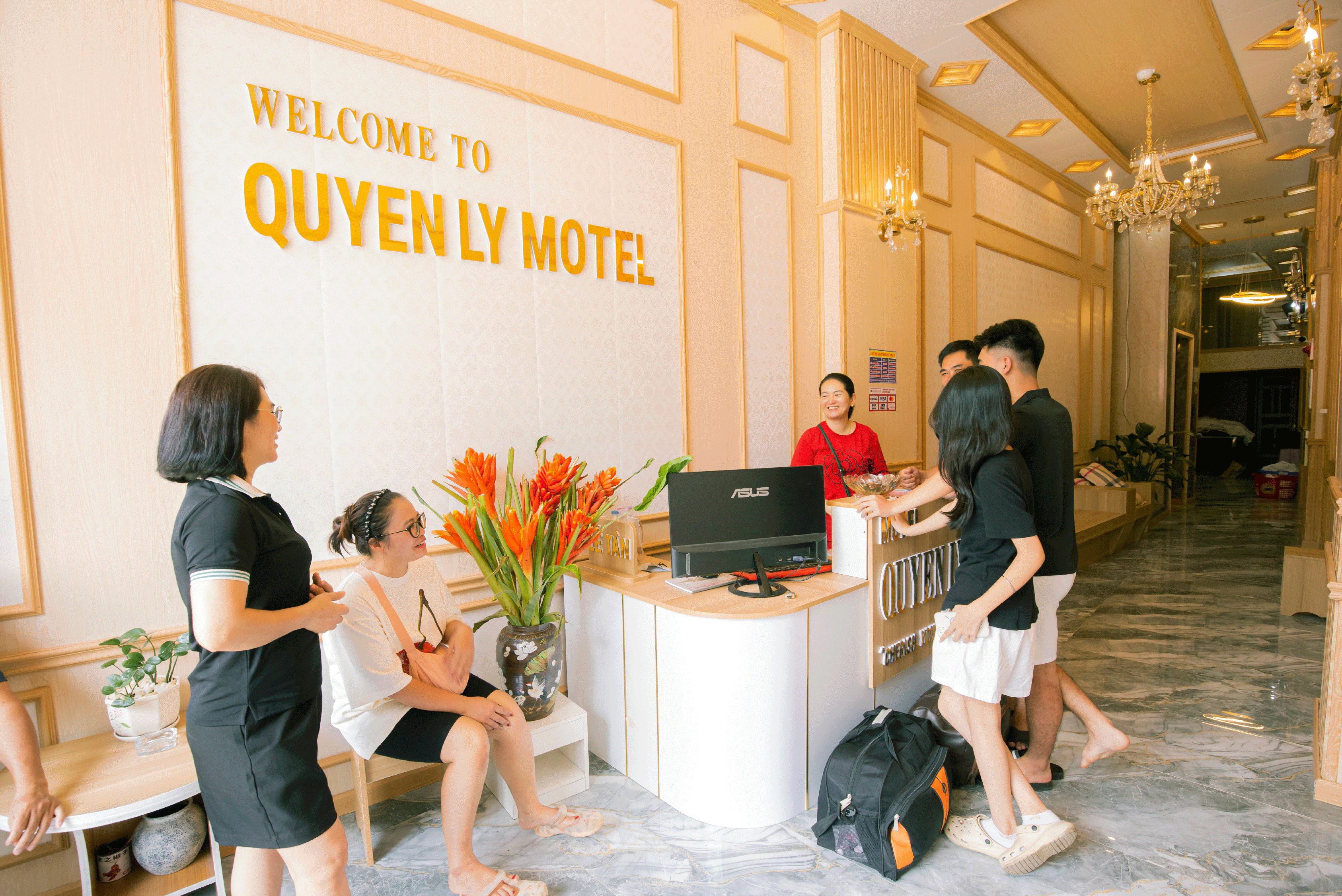 Una mesa de recepción con un cartel que dice 'WELCOME TO QUYENLY MOTEL', una computadora y un jarrón con flores.