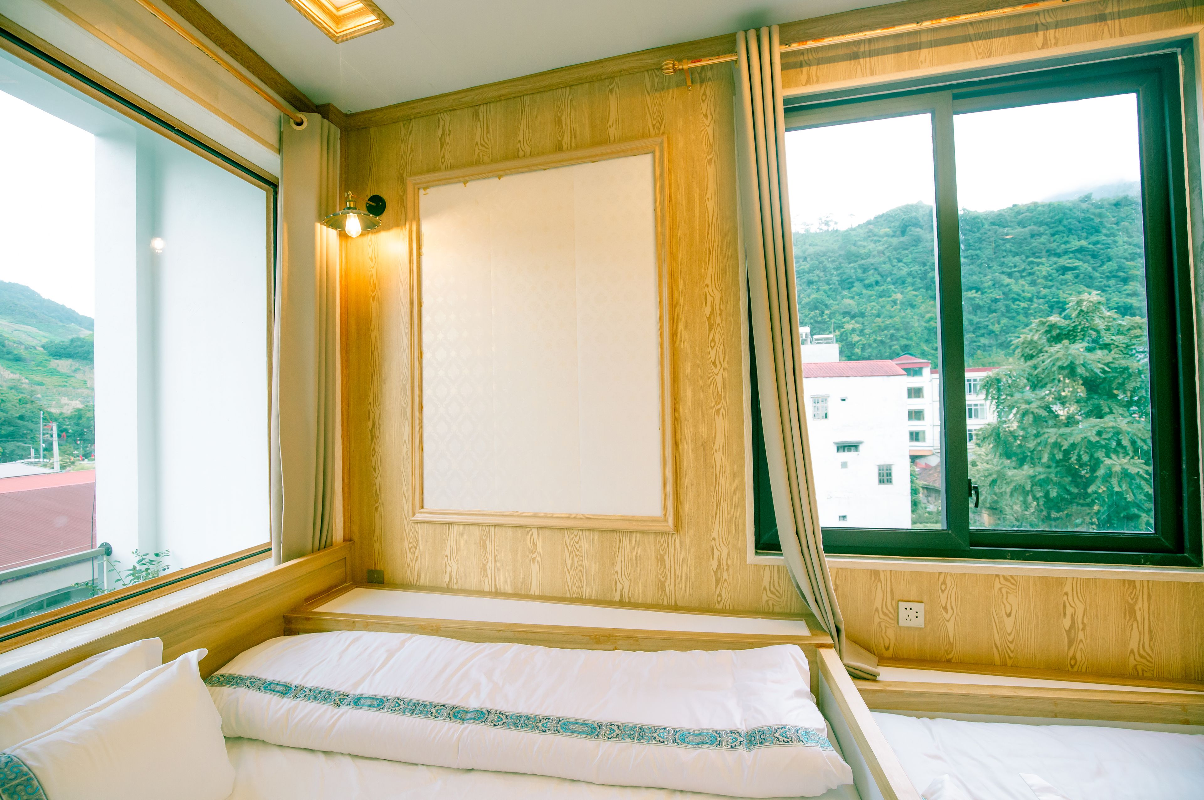 Une chambre avec un lit, une fenêtre avec des rideaux, un luminaire fixé au mur et une vue sur un immeuble et des collines.