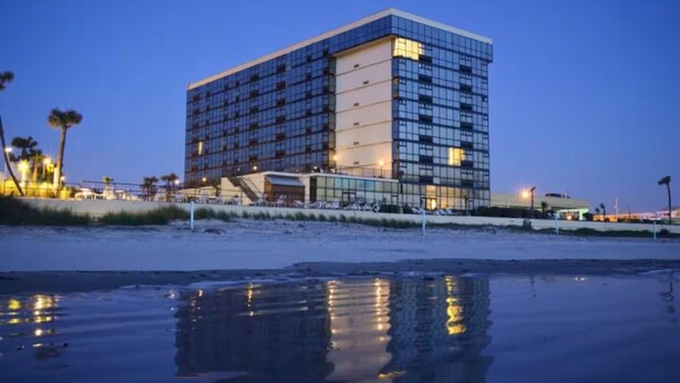 Un hôtel moderne à la façade en verre, situé près d’une plage dont l’eau a une surface réfléchissante.