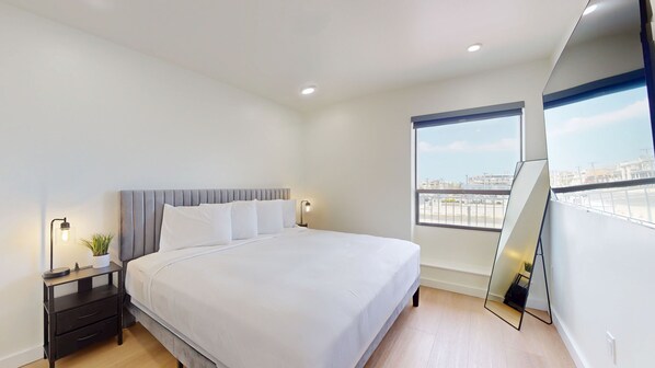 Een moderne slaapkamer met een groot bed, een nachtkastje met een lamp, een potplant en een groot raam met uitzicht op de stad.