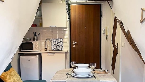 A compact kitchen with a microwave, sink, and small refrigerator. A dining table with plates and glasses is set up in front of a wooden door.