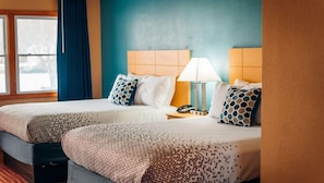 Quarto de hotel com duas camas, mesa de cabeceira com abajur e janela com cortinas.